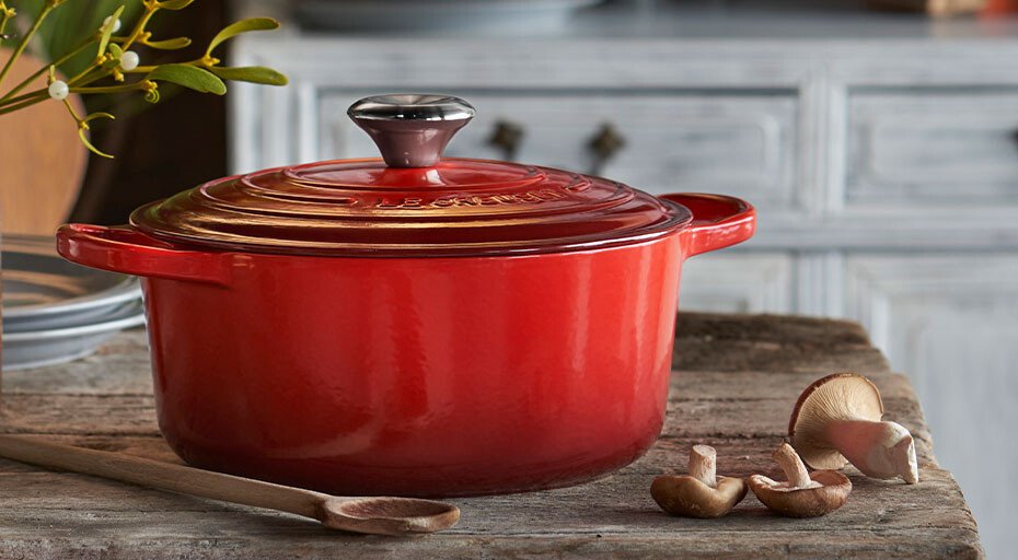 Roter Bräter von Le Creuset auf rustikalem Holztisch mit Holzlöffel und Pilzen, Hintergrund mit hellgrauem Schrank.