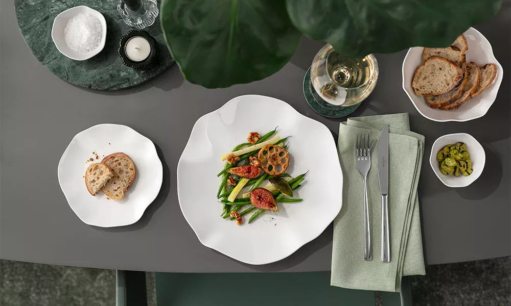 Eleganter Tisch mit Gemüsegericht, Brot, eingelegtem Gemüse, Glas Weißwein und Besteck, alles auf grünem Tischläufer.