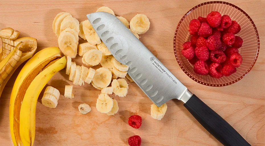 Santokumesser der Serie Partner von Wüsthof neben geschnittenen Bananen und Himbeeren auf Holzbrett, daneben rote Schale mit Himbeeren.