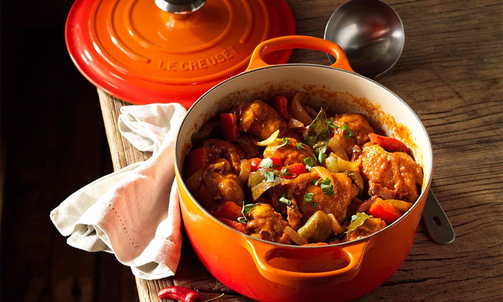 Oranger Bräter von Le Creuset mit geschmortem Hühnchen, Paprika und Zwiebeln in Tomatensauce auf Holzuntergrund, Löffel und Tuch daneben.