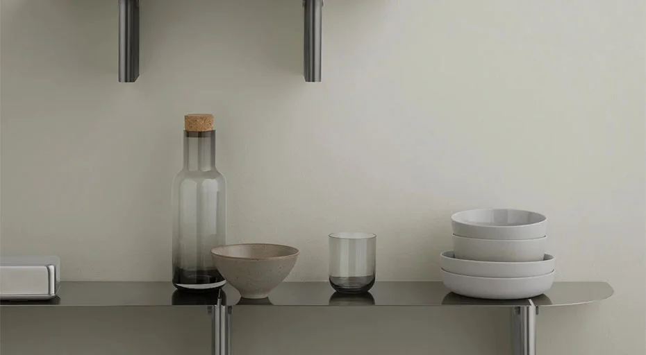 Regal mit Glasflasche, Tasse, Stapel Schüsseln und Glas auf grauer Oberfläche vor heller Wand.