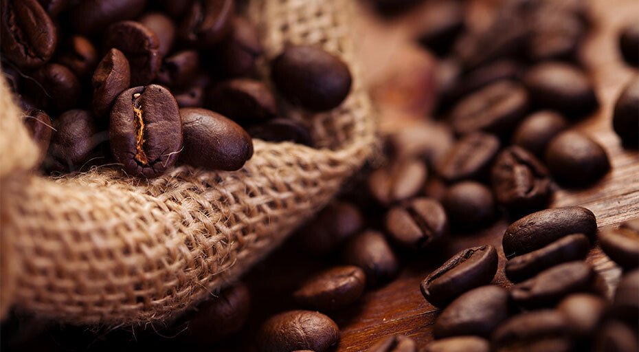 Nahaufnahme von Kaffeebohnen, die aus einem Jutesack auf eine Holzoberfläche fallen.