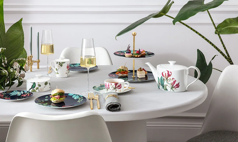 Eleganter Esstisch mit floralen Tellern, Glas Sektflöte, Teekanne, Burger, Kerzen und Pflanzen im Hintergrund.