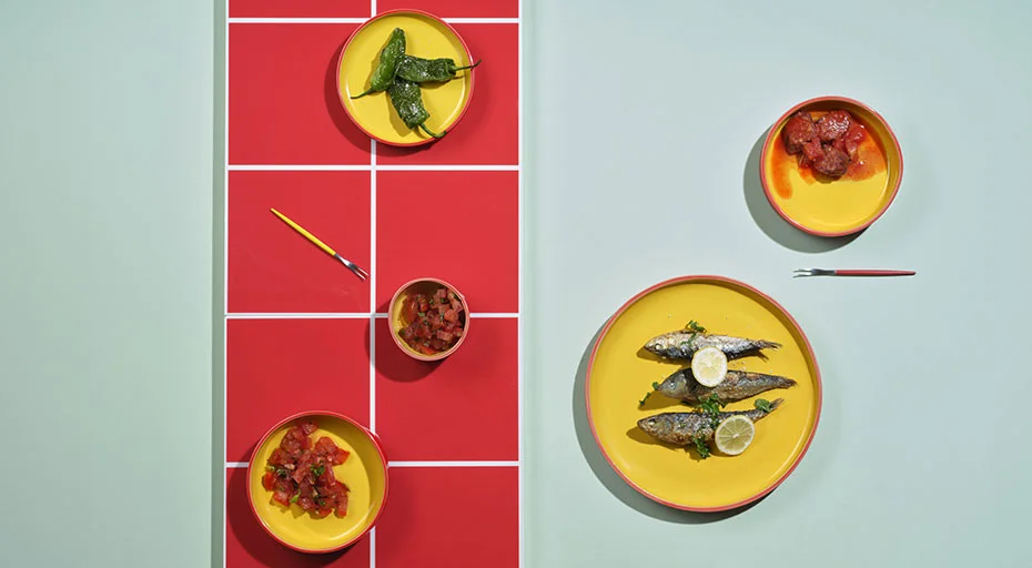 Gelbe Teller der Serie ASA Tapero Viva mit Sardinen, Zitronenscheiben, Paprika und Tomatenkompott auf rot-weißem Muster, minimalistisch arrangiert.