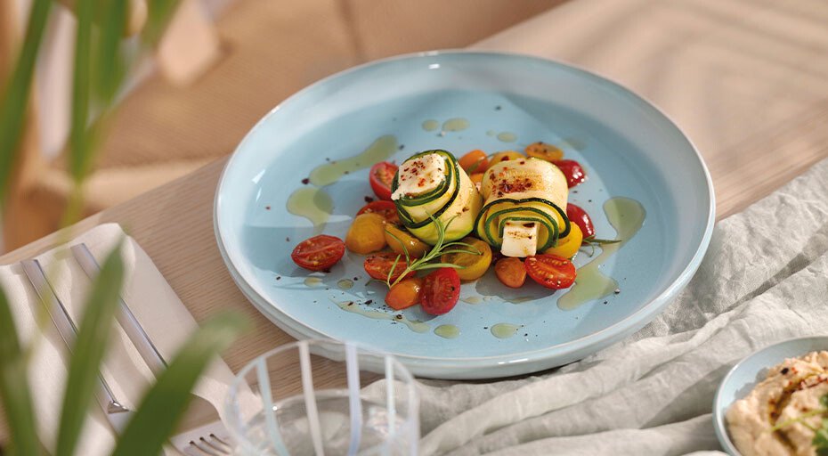 Kreative Zucchinirollen mit Feta auf Tomatensalat, serviert auf einem blauen Teller der Perlemor-Kollektion von Villeroy &amp; Boch, garnierte mit Olivenöl und Gewürzen.