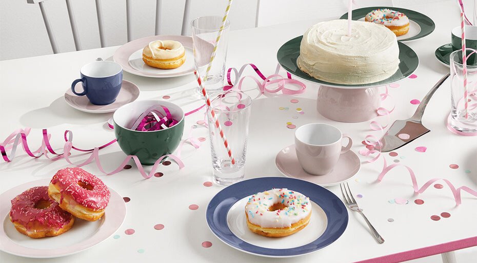 Feierlich gedeckter Tisch mit Donuts, Torte, Tassen, Gläsern und Dekorationen wie Luftschlangen und Konfetti.