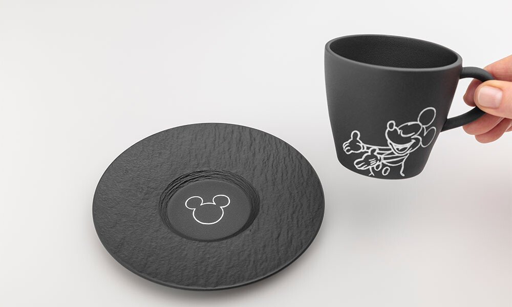 Schwarze Tasse und Untertasse der Serie Villeroy &amp; Boch Manufacture Rock – Mickey Mouse mit stilisierten Micky-Mouse-Motiven auf weißem Hintergrund, Tasse wird von einer Hand gehalten.