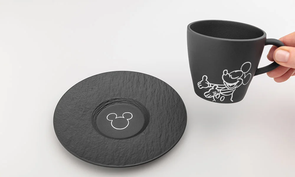 Schwarze Tasse und Untertasse der Serie Villeroy &amp; Boch Manufacture Rock – Mickey Mouse mit stilisierten Micky-Mouse-Motiven auf weißem Hintergrund, Tasse wird von einer Hand gehalten.