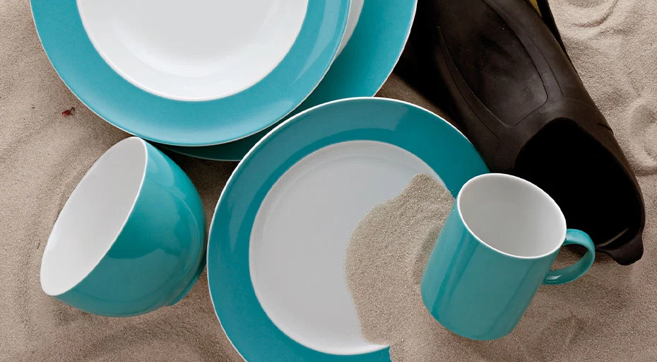 Türkises Geschirr der Serie Sunny Day Turquoise von Thomas und Flossen auf Sand angeordnet, mit teilweise bedecktem Teller und Tasse.
