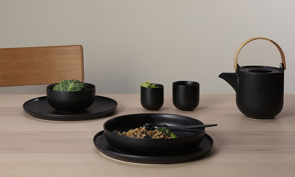 Minimalistische Tischszene mit schwarzem Geschirr, Teller, Schalen und Teekanne der Serie Coppa Kuro von ASA auf hellem Holztisch; modernes Design.