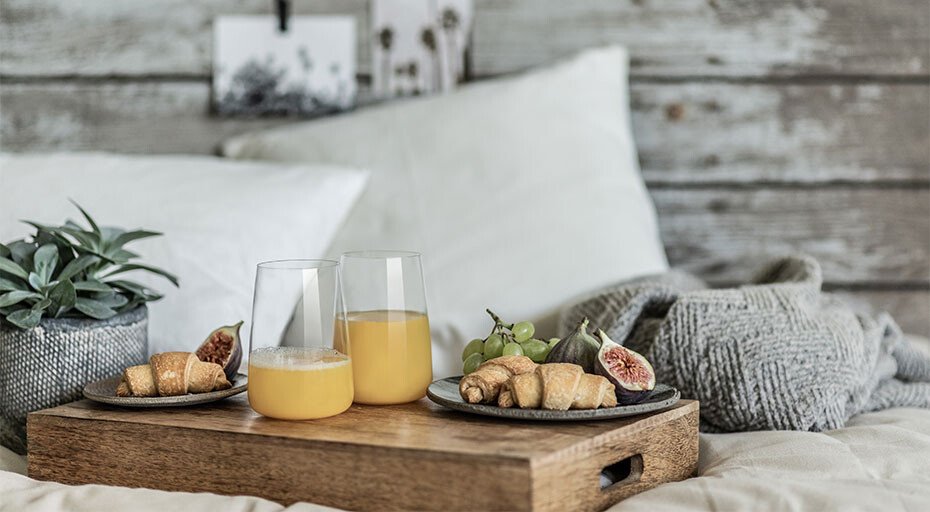 Frühstückstablett mit Orangensaft in Gläsern der Serie Vivid Senses von Zwiesel Glas, Croissants, Feigen und Trauben auf einem Bett, daneben eine Pflanze.