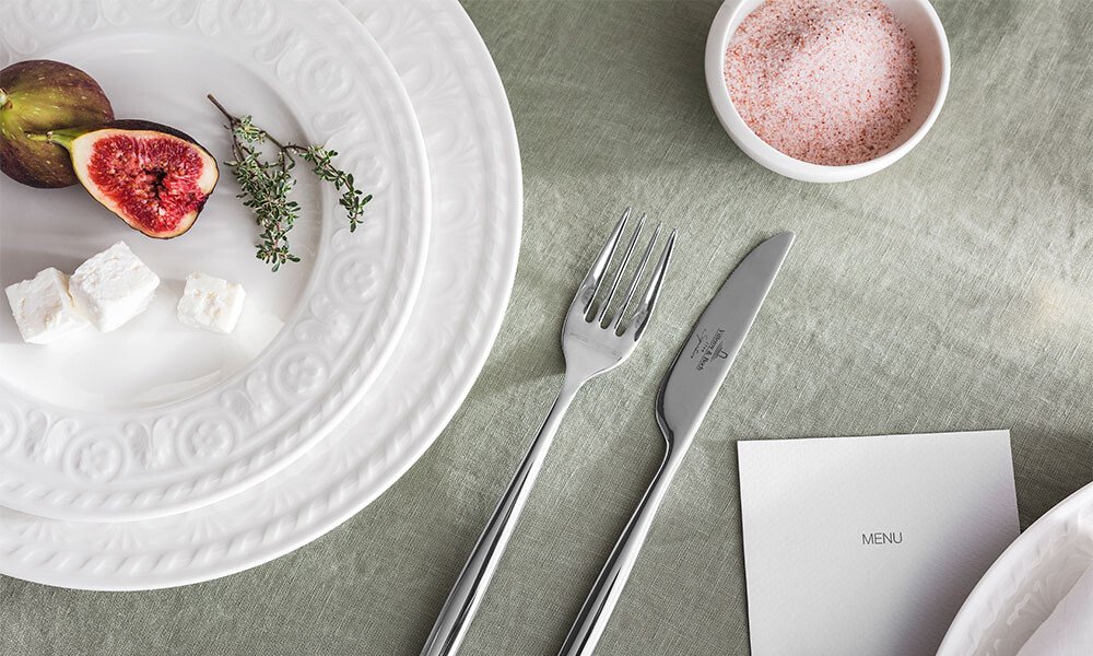 Weißer Teller der Serie Cellini von Villeroy &amp; Boch mit Feige, Thymian und Feta auf grünem Tischtuch, daneben Besteck und Schälchen mit rosa Salz.