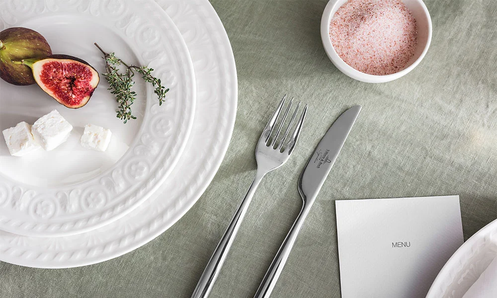 Weißer Teller der Serie Cellini von Villeroy &amp; Boch mit Feige, Thymian und Feta auf grünem Tischtuch, daneben Besteck und Schälchen mit rosa Salz.