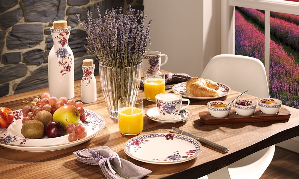 Mit Geschirr der Serie Artesano Lavendel Provencial von Villeroy &amp; Boch geschmackvoll gedeckter Tisch mit Obst, Croissants, Orangensaft und Lavendel in Vase, umgeben von malerischem Lavendelfeld.