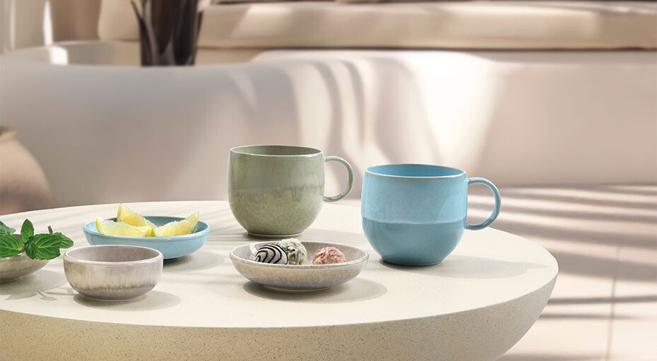 Heller Tisch mit zwei Tassen und drei Schälchen der Perlemor-Kollektion von Villeroy &amp; Boch, arrangiert mit Süßigkeiten und Zitronen, vor unscharfem, sonnigem Hintergrund.