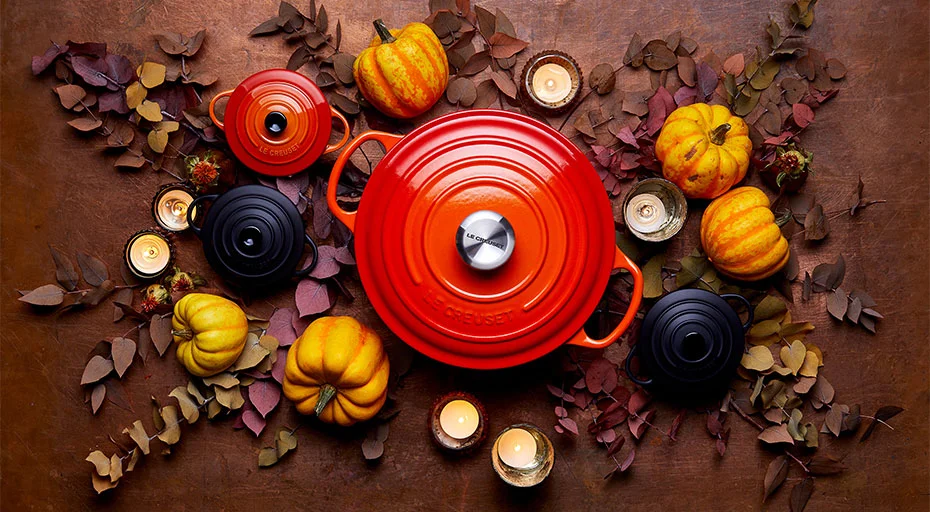 Roter Le Creuset Bräter, umgeben von Kürbissen, Herbstblättern und Kerzen auf braunem Untergrund angeordnet.