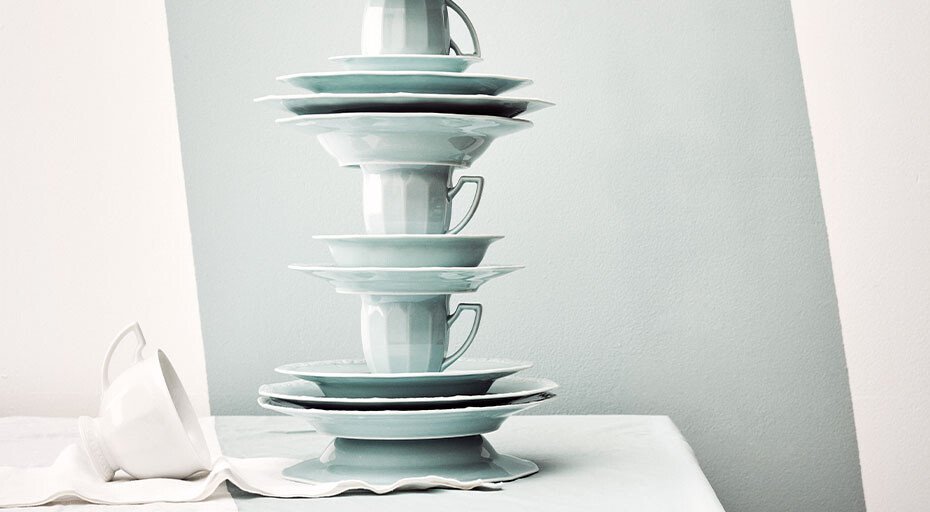Gestapelte, blaue Porzellanteller und Tassen der Serie Maria Pale Mint von Rosenthal auf weißem Tuch vor minimalistischem Hintergrund.