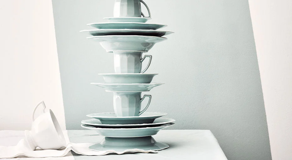 Gestapelte, blaue Porzellanteller und Tassen der Serie Maria Pale Mint von Rosenthal auf weißem Tuch vor minimalistischem Hintergrund.