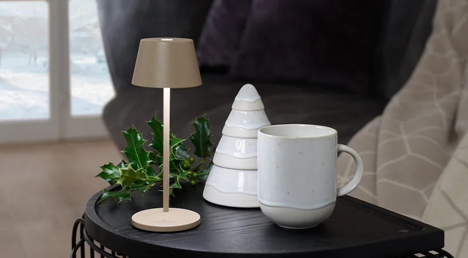 Tisch mit Lampe, Weihnachts-Tasse von Villeroy &amp; Boch, geometrischem Deko-Objekt und Stechpalmenzweig vor einem Fenster.