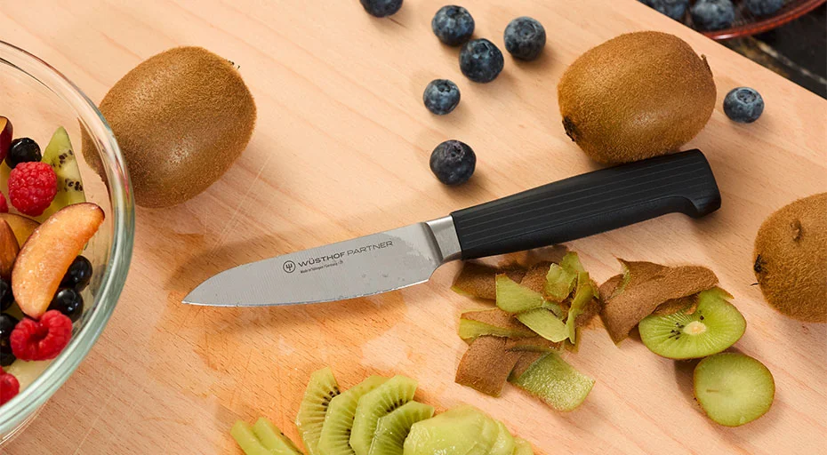 Ein schwarzes Messer der Serie Partner von Wüsthof auf einem Holzbrett neben geschälten und ungeschälten Kiwis, Blaubeeren und einer Obstschale.