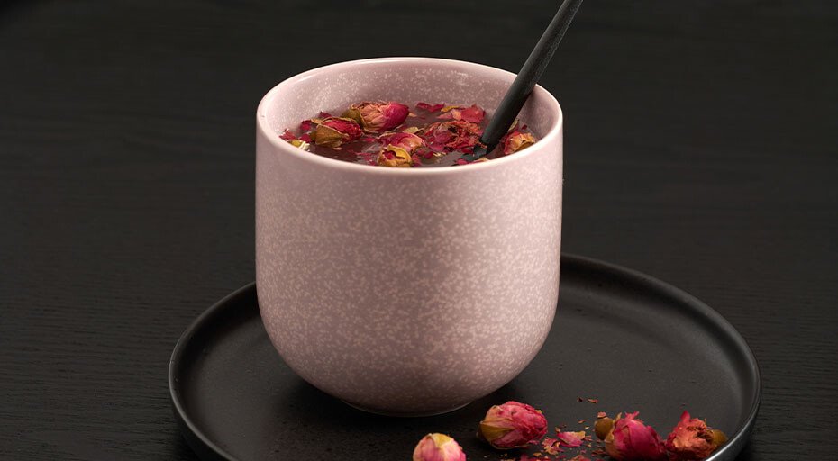 Ein rosa Becher der Serie Coppa Hanami von ASA mit Tee und Rosenblüten auf einem schwarzen Teller, schwarzer Hintergrund.