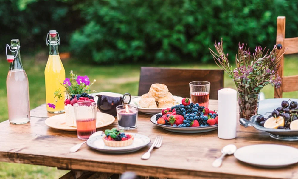 Gartenparty: Holztisch im Garten mit Getränken, Gebäck und Obst. Dekorativ mit Blumen und Lichterkette umrahmt.