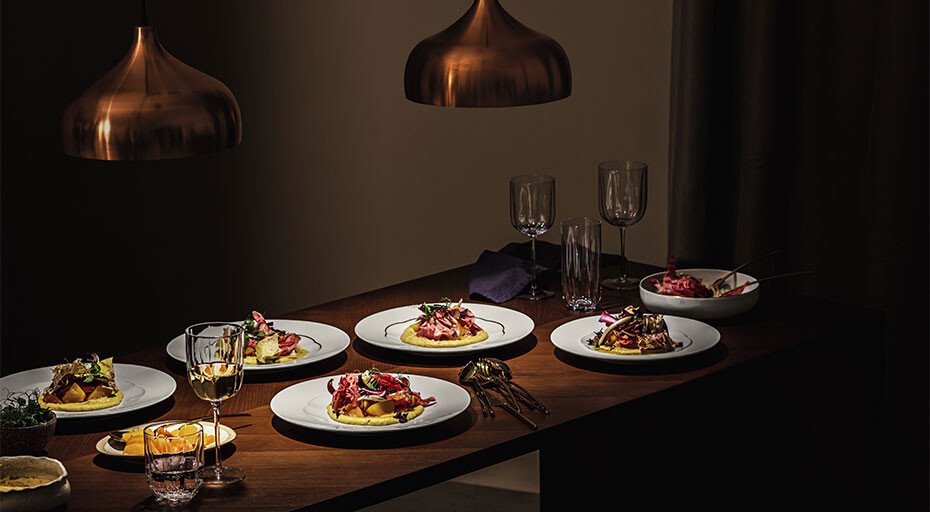 Eleganter Tisch mit sechs Gourmetgerichten, angerichtet auf Tellern der Serie Sonetto Oro von Rosenthal, beleuchtet von kupfernen Pendelleuchten, Weingläser und Dekoration.