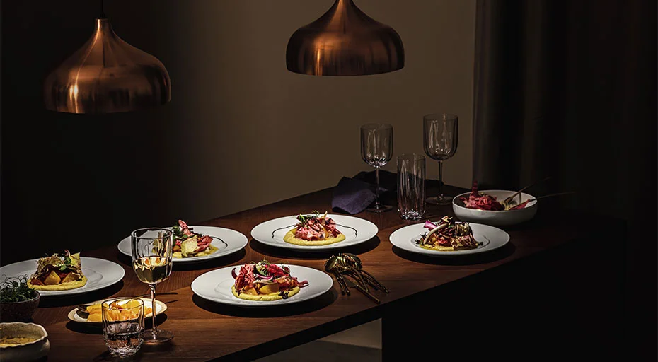 Eleganter Tisch mit sechs Gourmetgerichten, angerichtet auf Tellern der Serie Sonetto Oro von Rosenthal, beleuchtet von kupfernen Pendelleuchten, Weingläser und Dekoration.