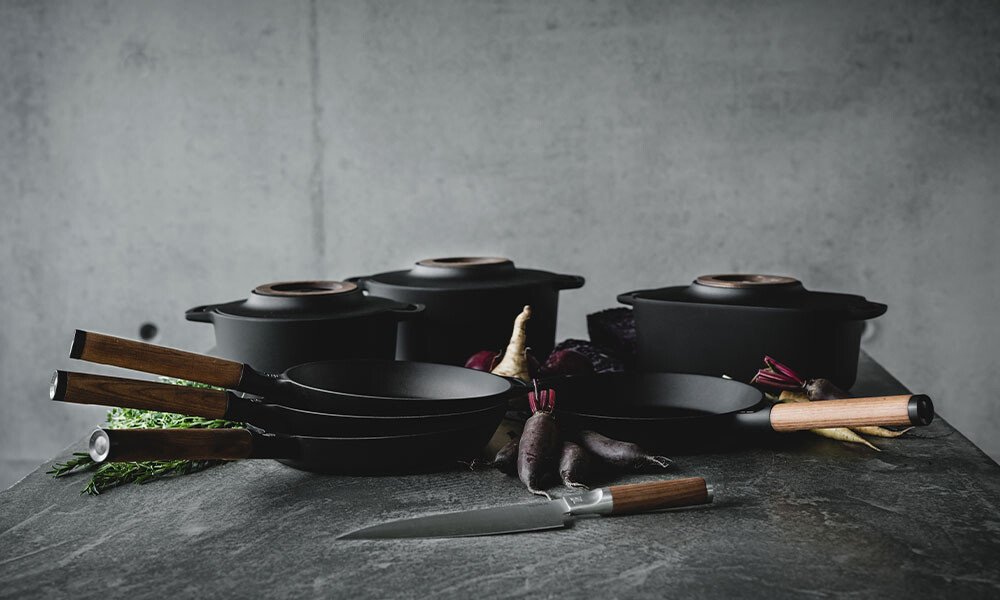 Kochgeschirr-Set aus Pfannen und Töpfen von Fiskars, auf grauem Untergrund stilvoll arrangiert, daneben Gemüse und ein Messer.