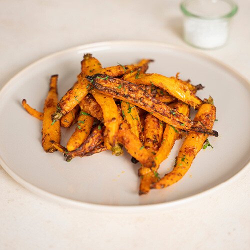 Karotten, gewürzt und angerichtet auf einem weißen Teller, im Hintergrund ein Glas mit weißem Inhalt.