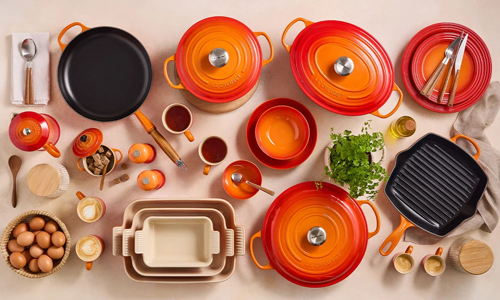 Verschiedene orangefarbene und rote Le Creuset Kochgeschirre, Töpfe und Formen, kunstvoll arrangiert auf einer Arbeitsfläche.