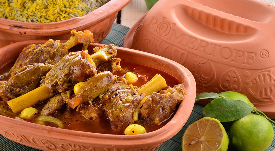 Lammkeulen in einem Römertopf mit Kochtopfdeckel, daneben Limetten und ein Topf mit Reis.