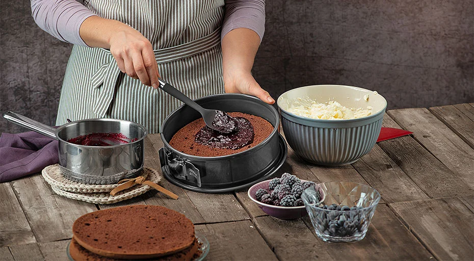 Person streicht Beerenfüllung auf Schokoladenboden in Kaiser-Springform, umgeben von Schüssel mit Creme, Beeren und Topf auf Holztisch.