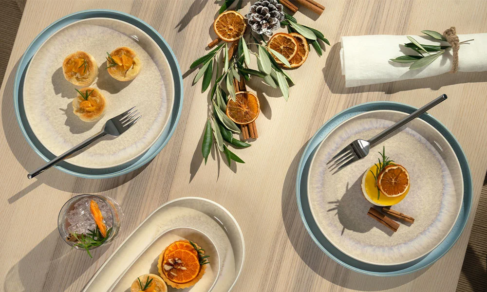 Tischarrangement mit Villeroy &amp; Boch Perlemor Tellern, Zimtstangen, getrockneten Orangenscheiben, Olivenzweigen und einem Getränkeglas.