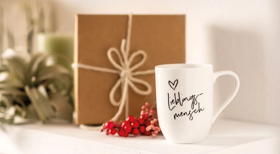Weiße Villeroy &amp; Boch Tasse mit Aufschrift &quot;Lieblingsmensch&quot; neben einem Geschenk und roten Beeren auf einem Regal.