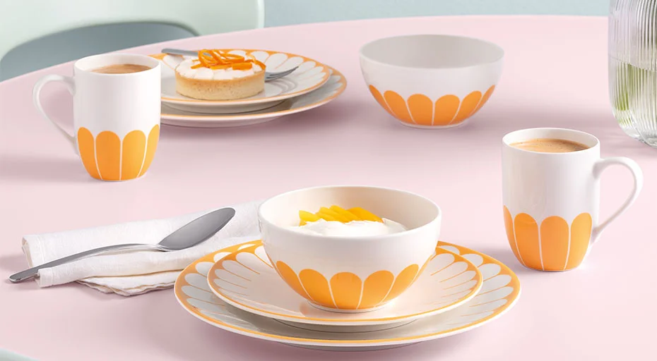 Frühstückstisch mit Gedeck von Villeroy &amp; Boch Fleur Soleil aus Tassen und Tellern in orange-weißem Muster, gefüllt mit Kaffee, Joghurt und Gebäck.