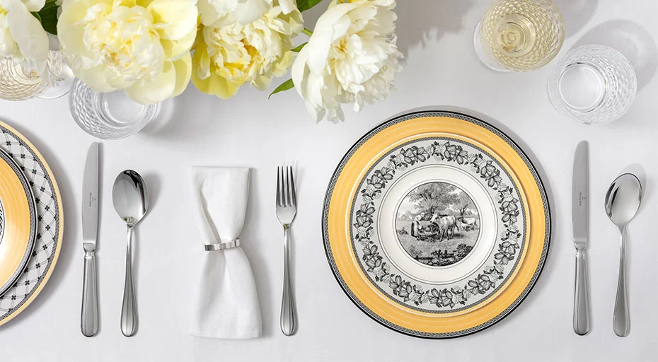 Elegantes Tischgedeck mit floralem Porzellanteller, Besteck und Kristallgläsern auf weißer Tischdecke, dekoriert mit gelben Blumen.