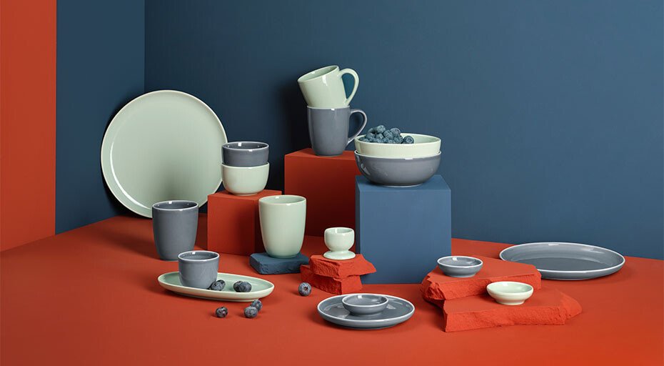 Keramikgeschirr in Grau, Weiß und Blau auf roten und blauen Podesten arrangiert, mit Blaubeeren auf rotem Untergrund.