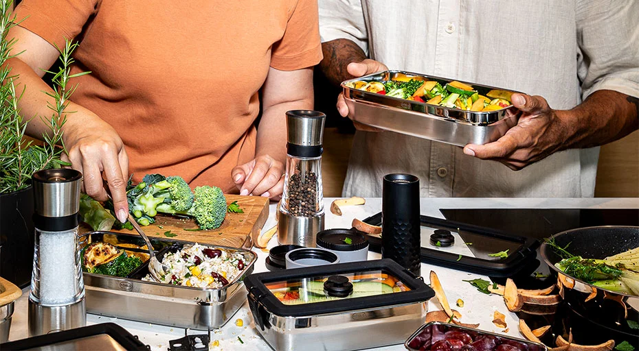 Zwei Personen bereiten Mahlzeiten in Edelstahl-Lunchboxen der Serie FLEXX 2GO von Rösle vor, umgeben von frischem Gemüse und Küchenutensilien.