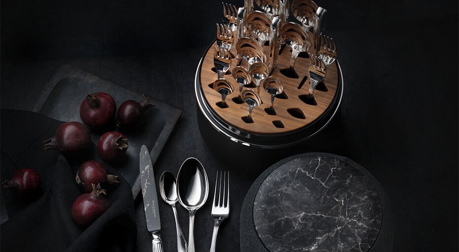 Ein schwarzes Tischset mit Besteckständer The Box von Robbe &amp; Berking, Tellern, Löffeln, Gabeln, Messer und roten Zwiebeln auf dunklem Untergrund.