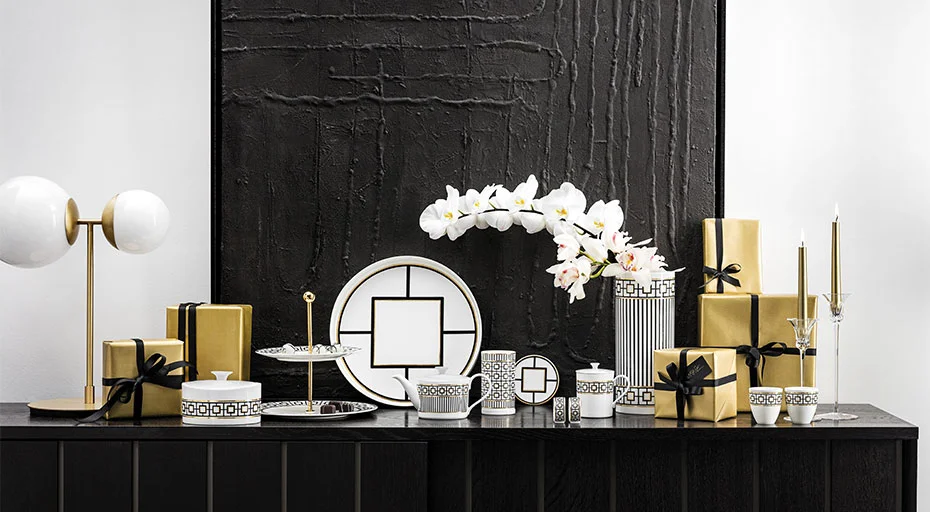 Elegantes Tischarrangement mit Villeroy &amp; Boch MetroChic Porzellangeschirr, goldenen Geschenkboxen und Orchideen vor schwarzer Texturwand.