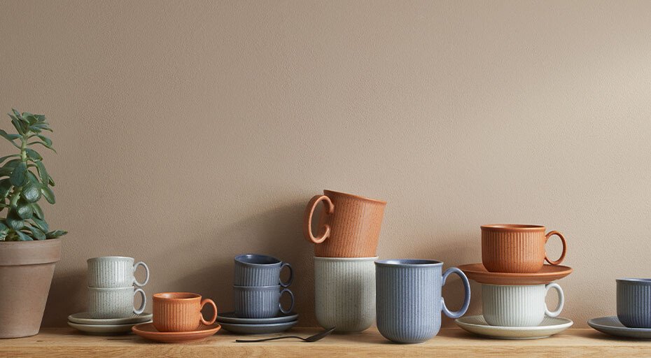 Keramikgeschirr der Serie Clay von Thomas in Erdtönen, unterschiedlicher Höhe, auf einem Holztisch vor beiger Wand. Links steht eine grüne Topfpflanze.