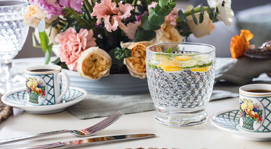 Gedeckter Tisch mit Blumengesteck, zwei verzierte Kaffeetassen und einem Kristallglas von Villeroy & Boch, befüllt mit Wasser und Zitronenscheiben.