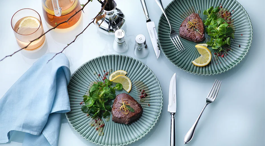 Zwei Teller mit Steak, Salat, Zitrone. Besteck von Robbe &amp; Berking, Getränke und blaue Serviette auf hellem Tisch arrangiert.