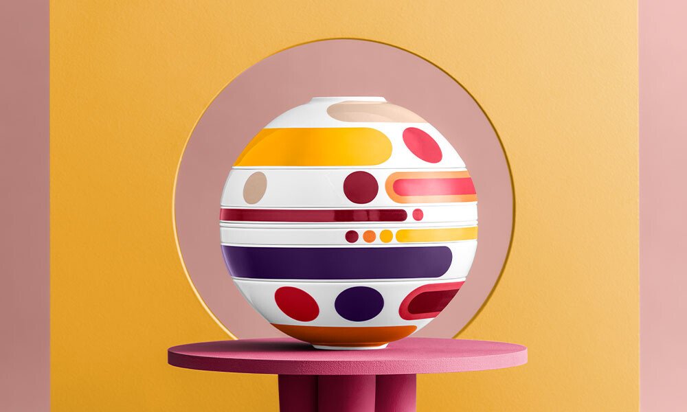 La Boule Geschirr-Set von Villeroy &amp; Boch mit buntem, geometrischem Muster vor gelbem und rosa Hintergrund, platziert auf rundem, violettem Tisch.