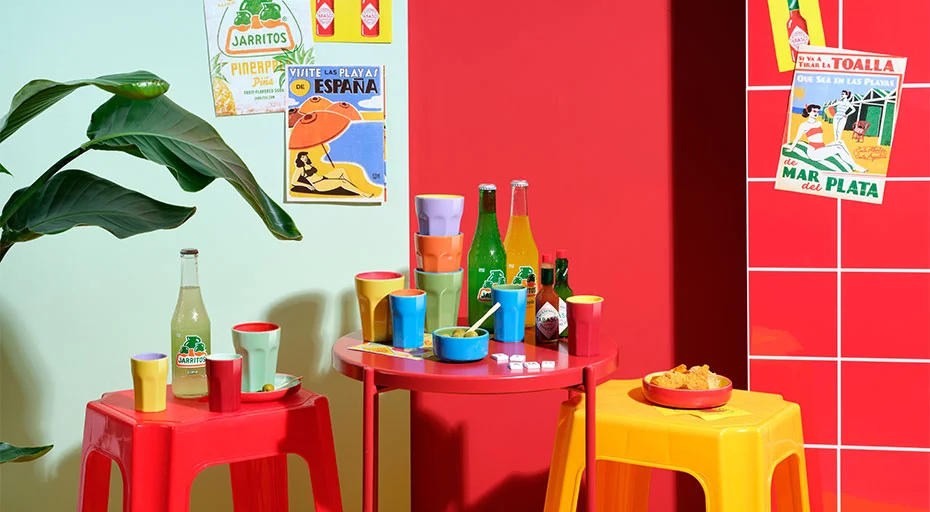 Buntes ASA Tapero Viva Geschirr mit Getränke und Snacks auf roten und gelben Hockern, umgeben von Retro-Postern und einer Pflanze vor rot-grüner Wand.
