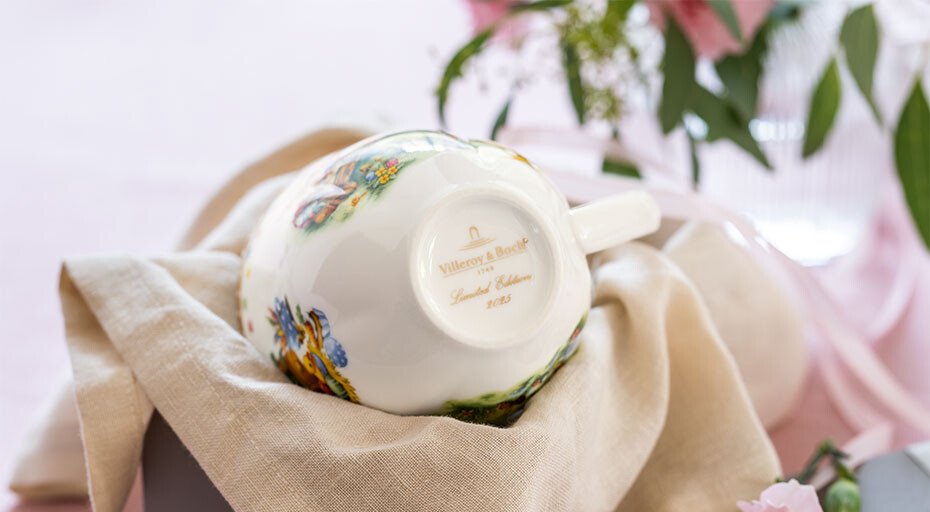 Tasse der Annual Easter Edition von Villeroy &amp; Boch mit floralem Muster, liegend auf einem Tuch, umgeben von Blumenarrangement.