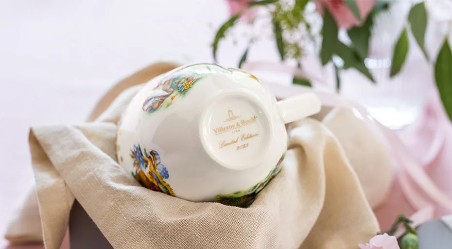 Tasse der Annual Easter Edition von Villeroy &amp; Boch mit floralem Muster, liegend auf einem Tuch, umgeben von Blumenarrangement.