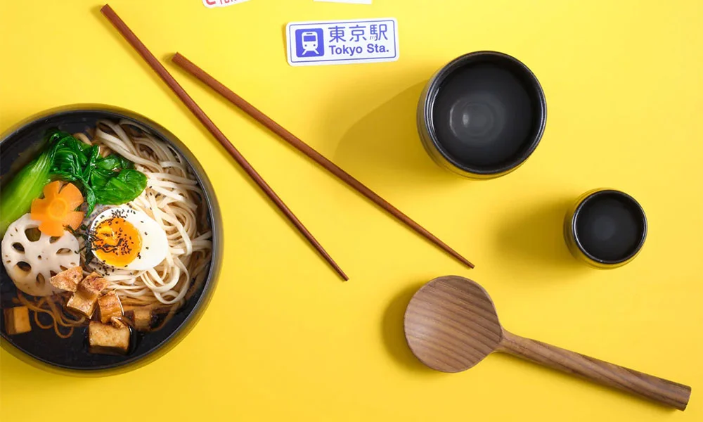 Schüssel mit Ramen, Eihälfte, Gemüse und Tofu auf gelbem Hintergrund, daneben Essstäbchen und Schöpflöffel.
