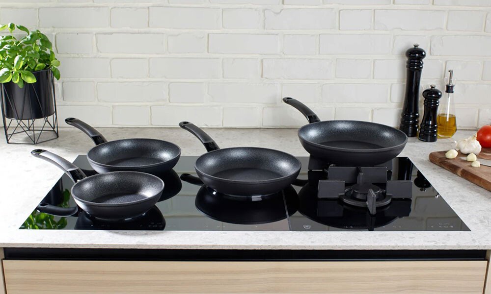 Drei schwarze Pfannen und ein Wok der Serie Adamant von Fissler auf einem Glaskeramik-Kochfeld vor weißer Backsteinwand und Gewürzen.